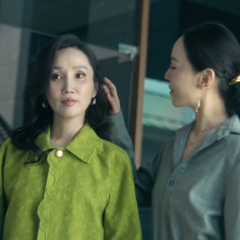Park Bo-kyung de jaqueta verde, no papel de Jeong Yeo-jin, ajeitando o cabelo diante do espelho, com a Sarah Kim de Shin Hae-sun atrás dela, em A Arte de Sarah