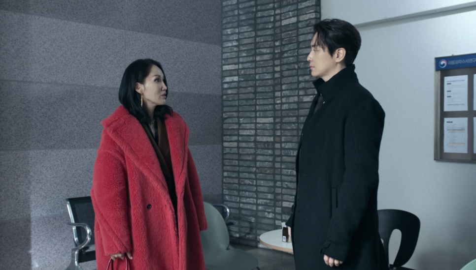 A Jeong Yeo-jin de casaco vermelho conversando de pé com o detetive Park Mu-gyeong, de sobretudo preto, numa sala da delegacia