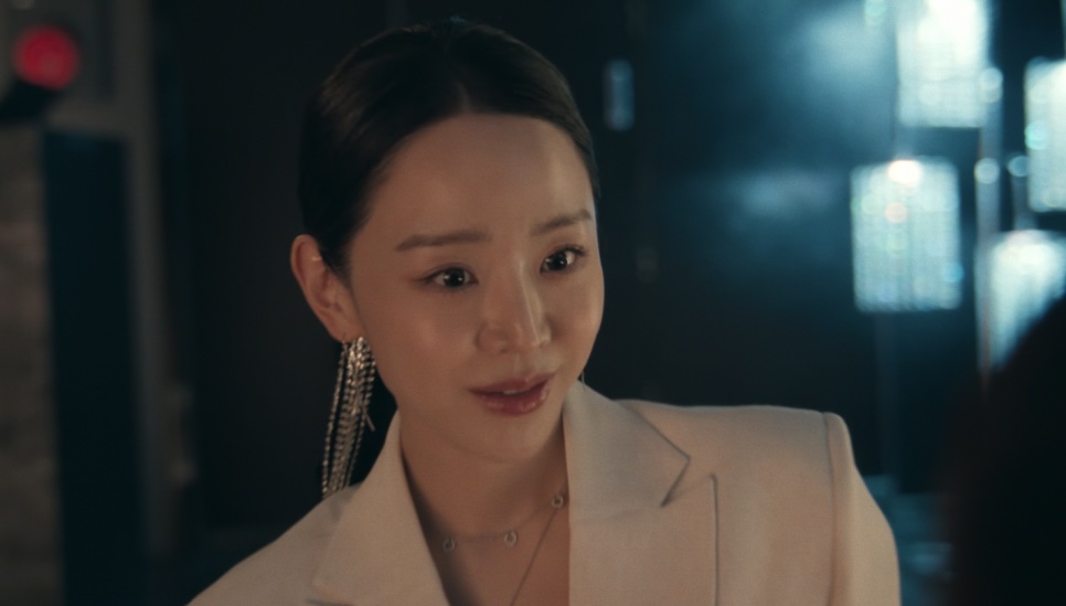 Shin Hae-sun de blazer claro e brincos longos, no papel de Sarah Kim, sorrindo num bar