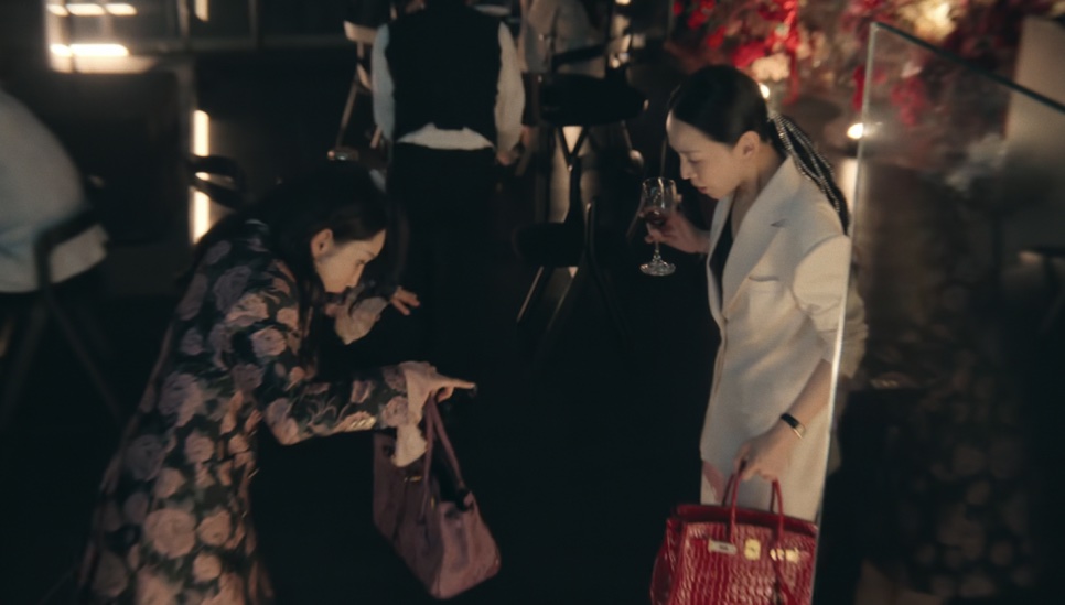 A Jeong Yeo-jin se abaixando para olhar a bolsa dela, enquanto a Sarah Kim segura uma taça de vinho e uma bolsa Birkin vermelha