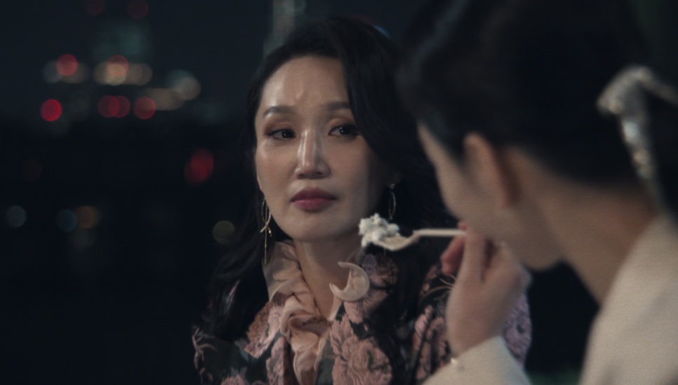 A Jeong Yeo-jin dando uma colherada de sobremesa na boca da Sarah Kim, com as luzes da cidade ao fundo