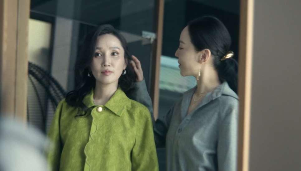 A Jeong Yeo-jin de jaqueta verde diante do espelho, com a Sarah Kim de camisa cinza atrás dela