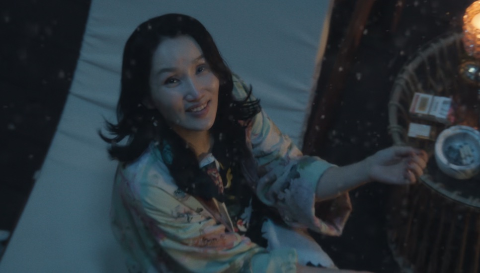 A Jeong Yeo-jin de roupão estampado, sentada numa varanda à noite, estendendo a mão até uma mesinha