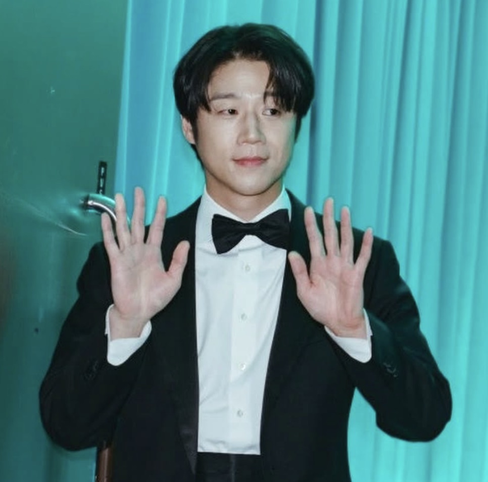 Jung Joon-won de smoking, com as duas mãos levantadas, diante de uma cortina verde