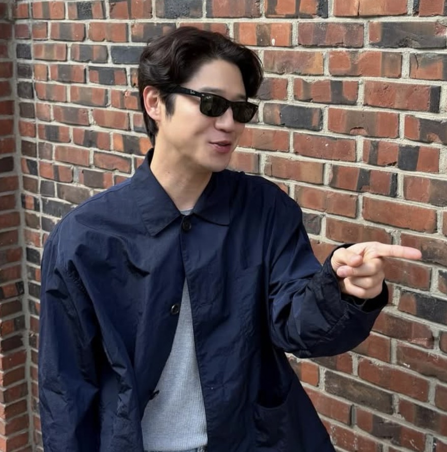 Jung Joon-won de óculos escuros e jaqueta azul-marinho, diante de uma parede de tijolos