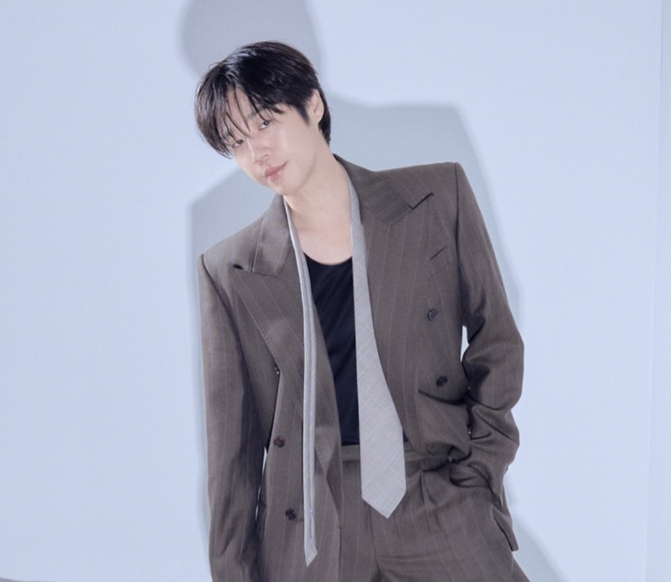 Jung Joon-won de terno marrom e gravata clara, diante de um fundo azul-claro
