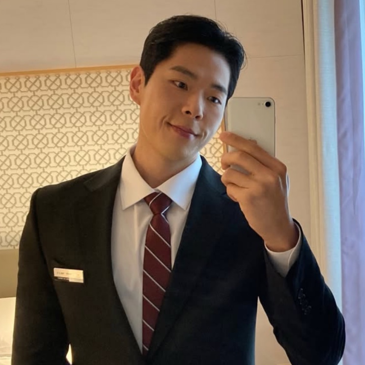 Joo Yeon-woo de terno escuro e gravata vinho, tirando uma selfie no espelho