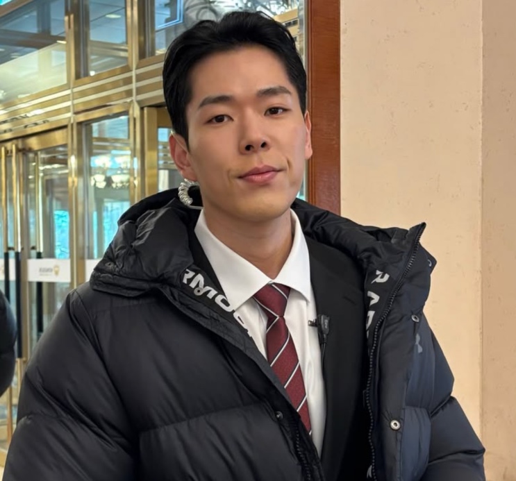 Joo Yeon-woo de terno com jaqueta preta por cima, na entrada de um prédio