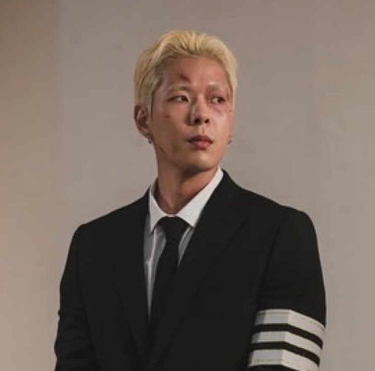 Joo Yeon-woo de cabelo platinado e terno preto com faixas brancas na manga