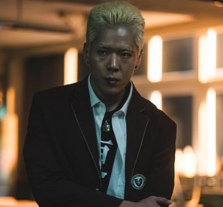 Kim Soon-cheol de cabelo platinado e uniforme escolar, de braços cruzados