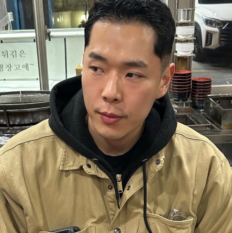 Joo Yeon-woo de jaqueta bege e moletom preto, sentado em um restaurante