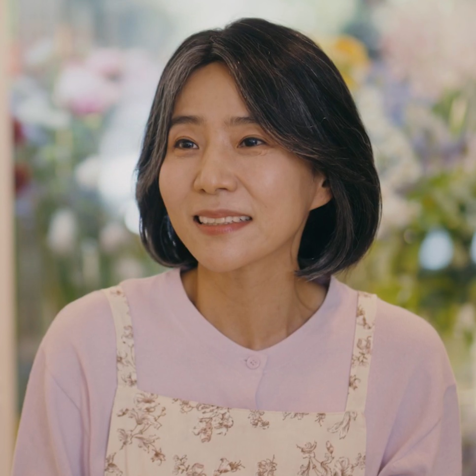 A Bae Yoon-ok sorrindo, de avental florido, com flores ao fundo