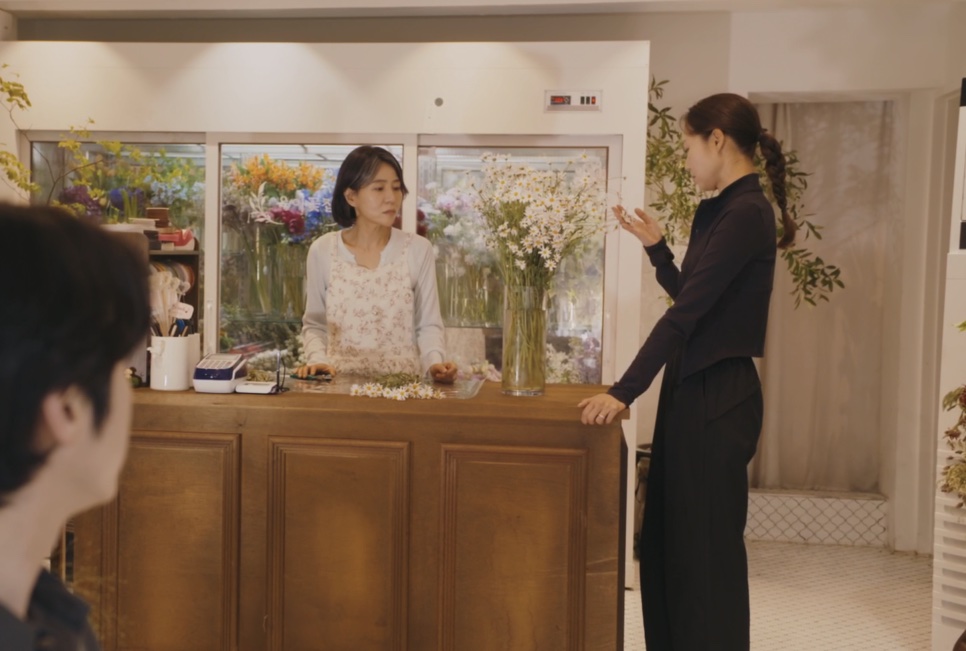 A Bae Yoon-ok atrás do balcão da floricultura, conversando com uma cliente