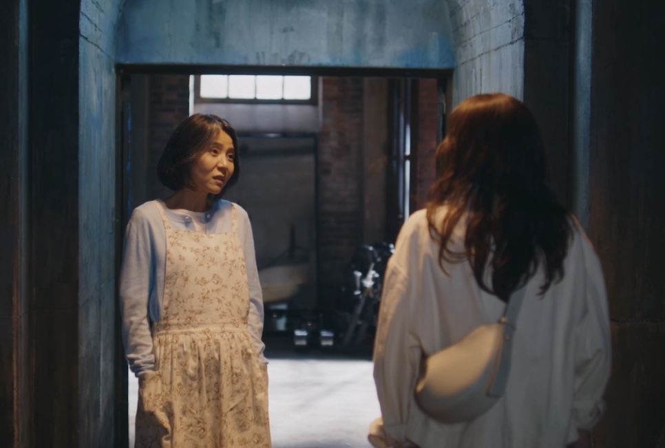 A Bae Yoon-ok de avental, parada numa porta, olhando para uma mulher de costas