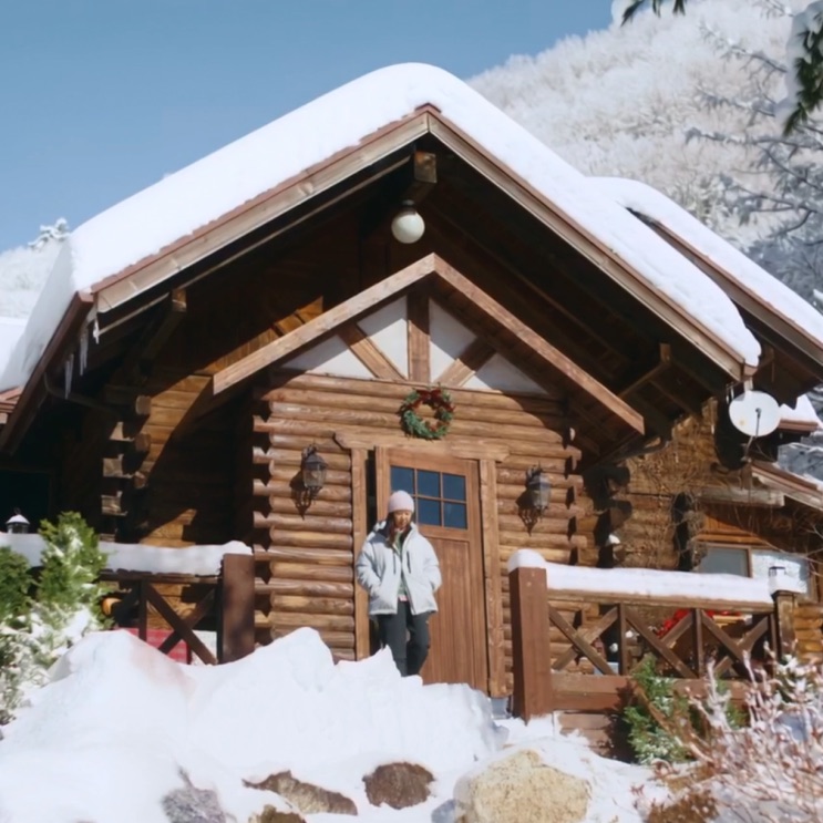 Cabana de troncos coberta de neve, com uma pessoa de casaco claro parada na porta