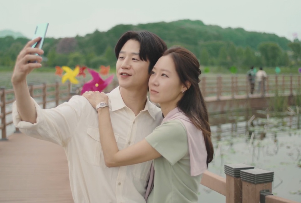 Kwon Tae-seong tirando uma selfie abraçado com Yu Bo-na no deck de madeira do parque