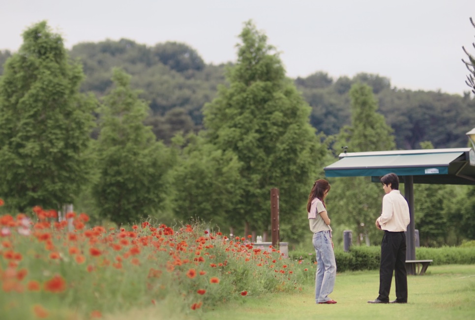 Yu Bo-na e Kwon Tae-seong conversando num gramado, ao lado de um canteiro de papoulas vermelhas