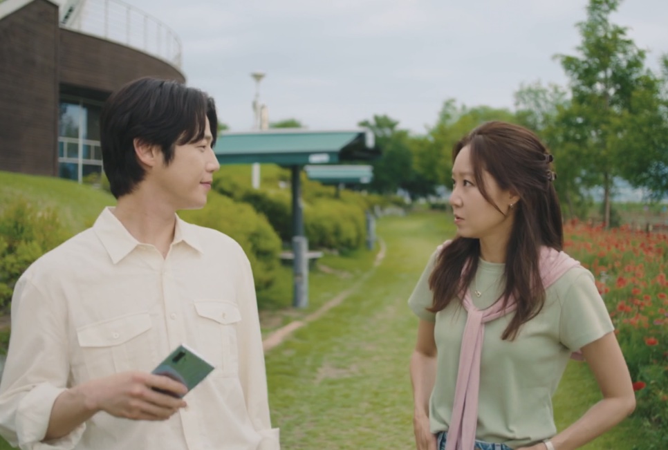 Kwon Tae-seong, com o celular na mão, e Yu Bo-na se olhando num caminho de grama do parque