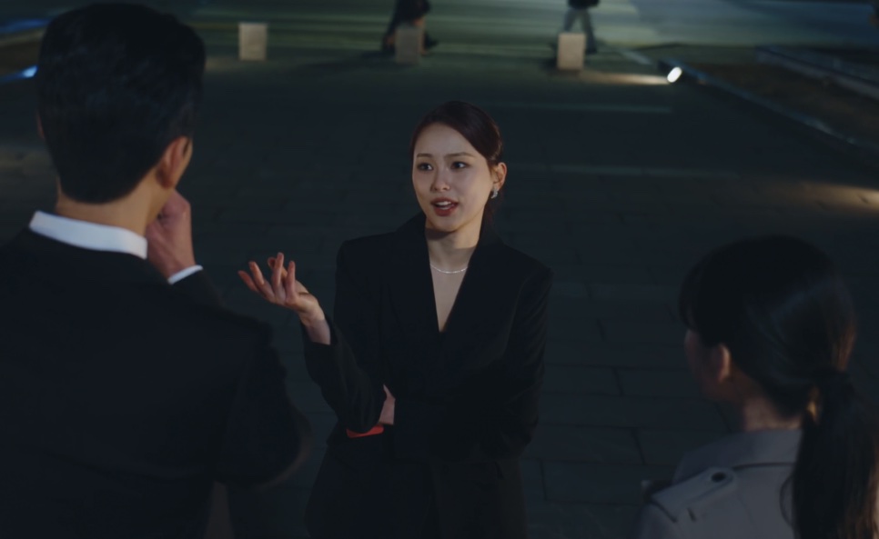 Mulher de terno preto conversando com duas pessoas de sobretudo, à noite, em cena de A Bona Fide Killer