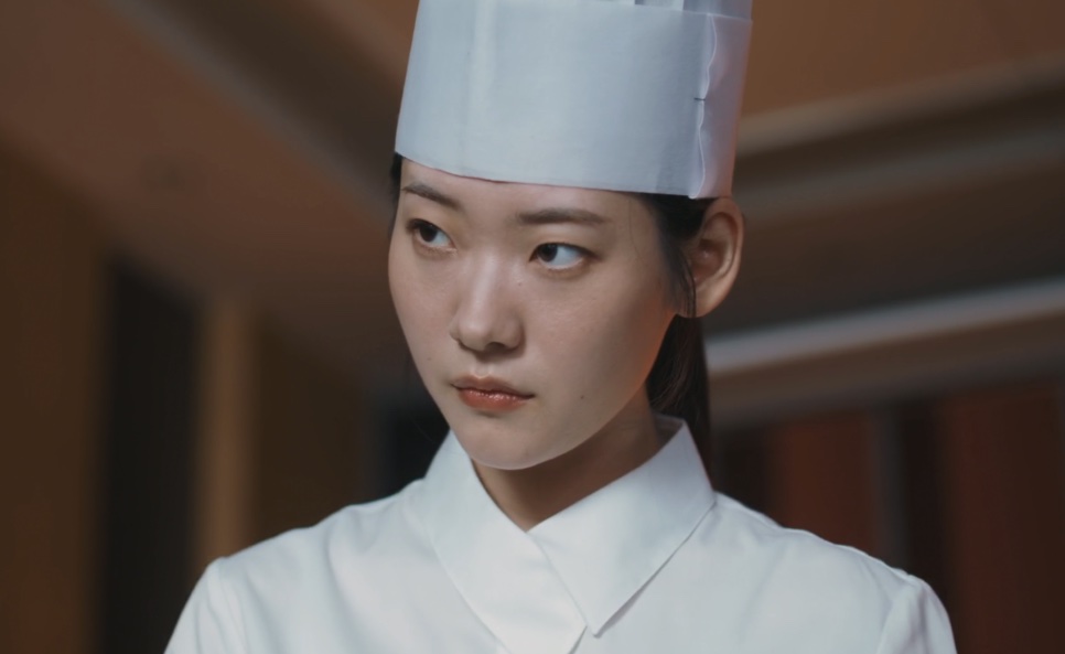 Moon Meo-ru de uniforme branco de cozinha e touca, olhando de lado no corredor do hotel