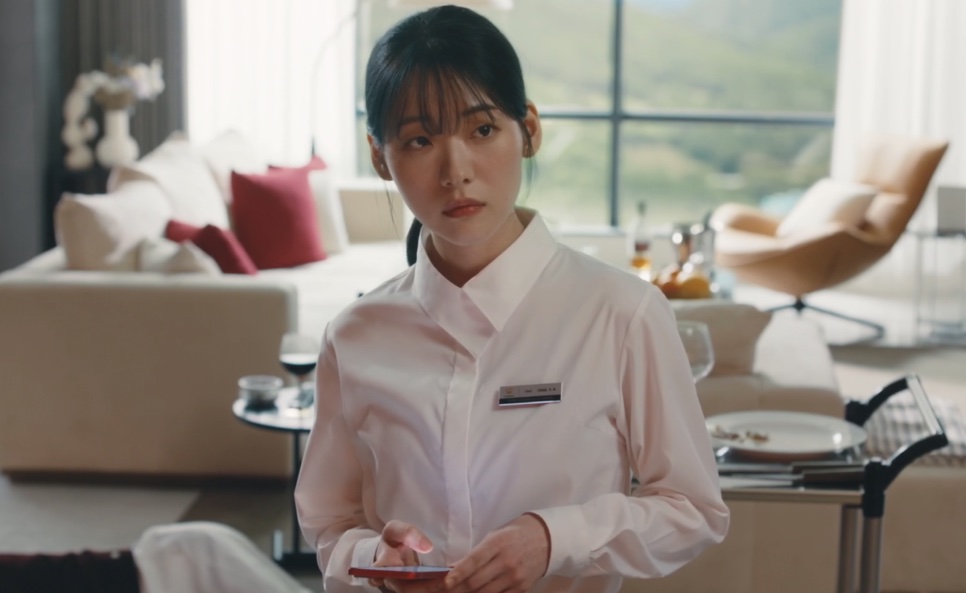 Moon Meo-ru de camisa branca e crachá, em pé dentro de uma suíte do hotel