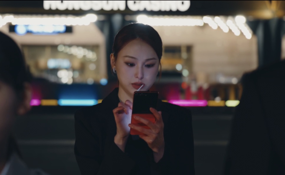 Moon Da-rae mexendo no celular em frente à fachada iluminada do Hongsun Casino