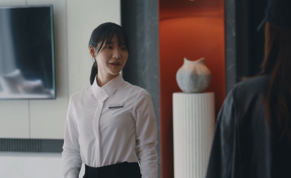 Moon Meo-ru de camisa branca, sorrindo de leve para alguém dentro da suíte do hotel