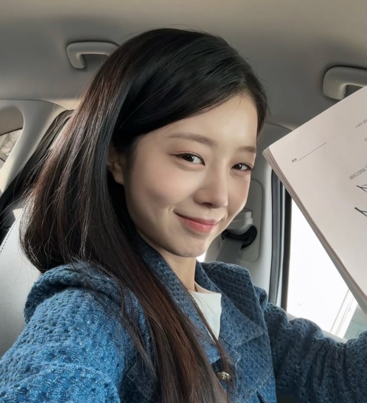 Han Chae-rin de casaco azul, em selfie dentro do carro, com o roteiro ao lado