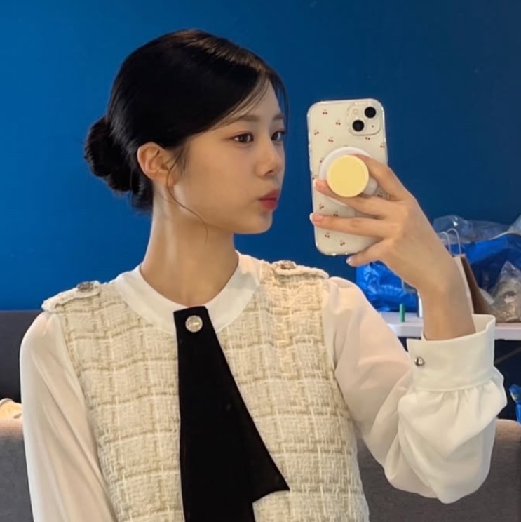 Han Chae-rin de coque, fotografando o próprio reflexo no espelho, de blusa branca e colete de tweed