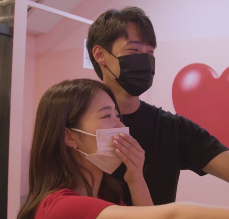 Han Chae-rin e um participante do reality Some Ping, os dois de máscara, numa sala rosa