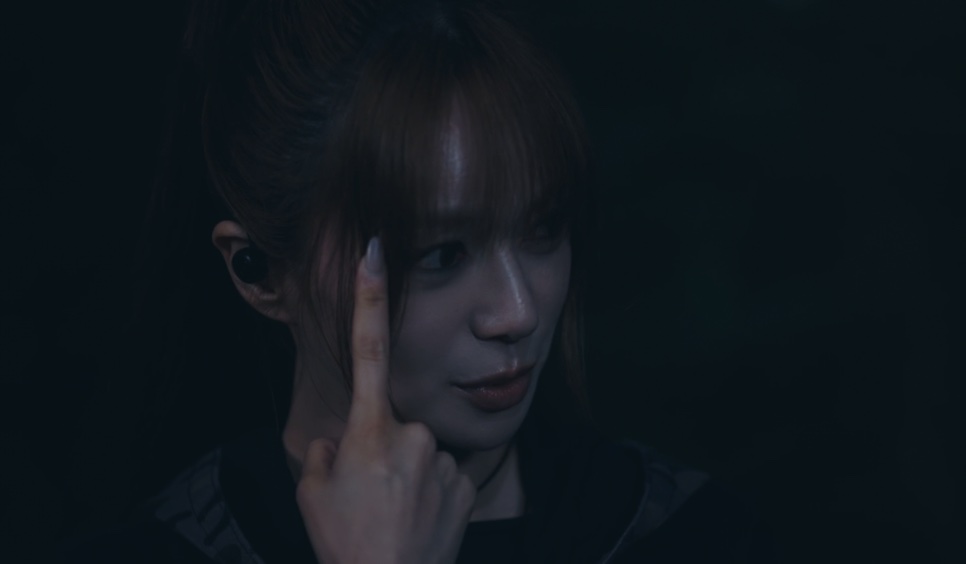 A Layla, personagem da Lee Elijah, com o dedo no fone de ouvido, numa cena noturna