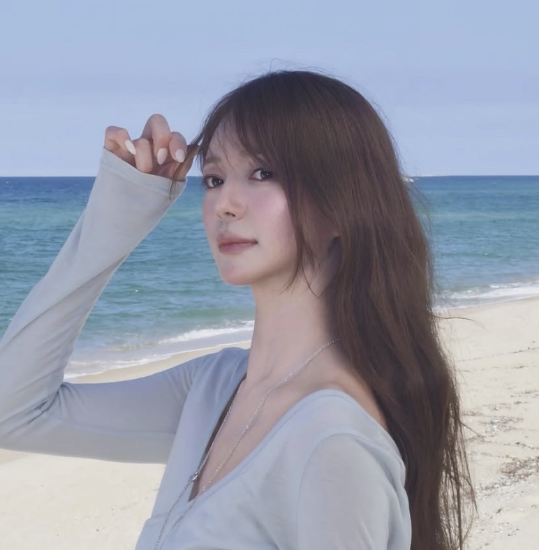 Lee Elijah de blusa azul-clara, com a mão no cabelo, numa praia