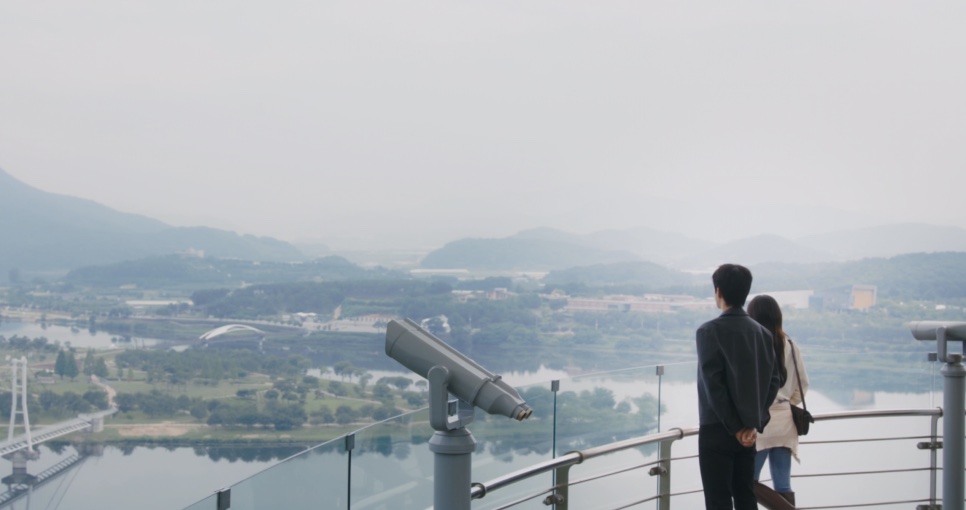 O casal de costas no mirante, ao lado de uma luneta, olhando o rio e uma ponte suspensa
