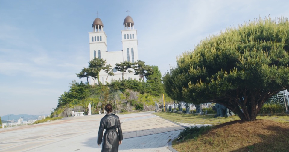 Yu Bo-na de casaco de couro preto caminhando em direção a uma igreja branca de duas torres