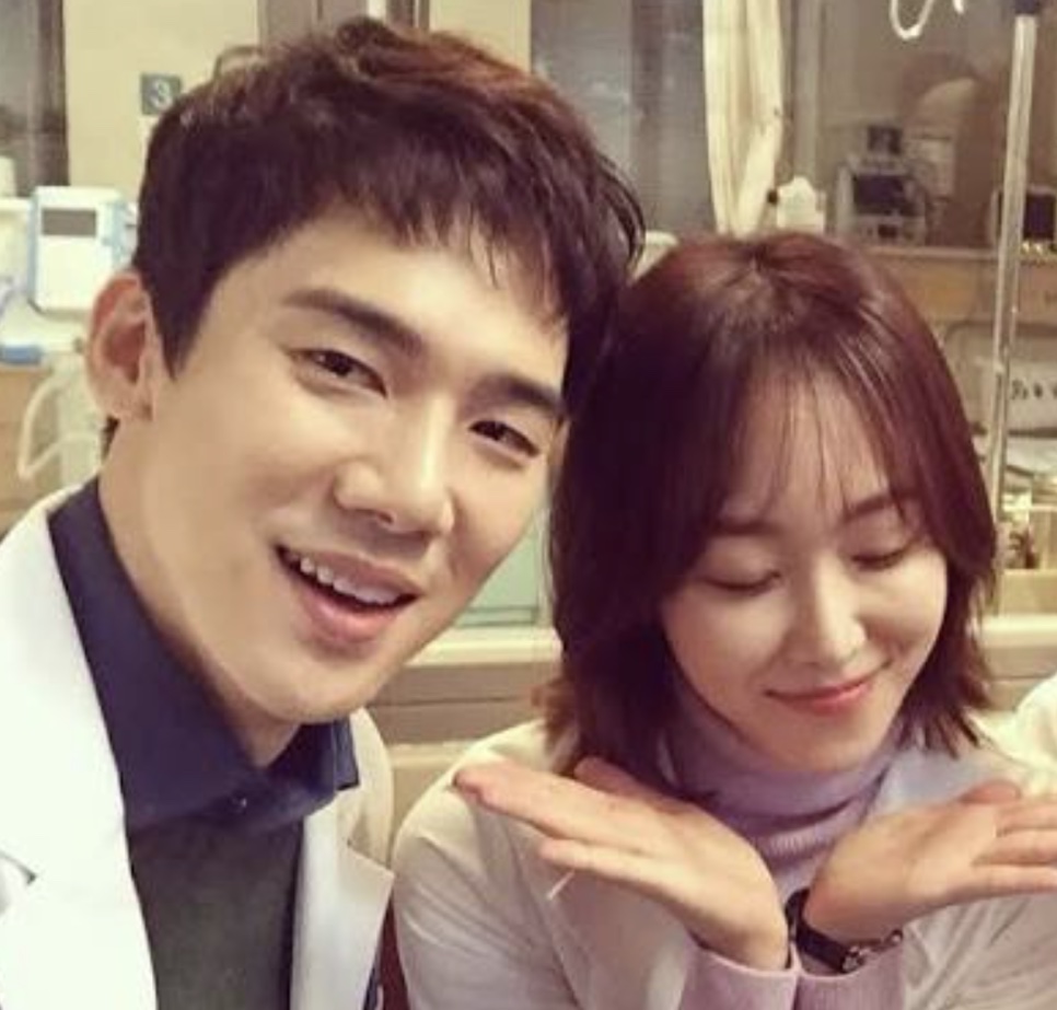 Yoo Yeon-seok e Seo Hyun-jin sorrindo lado a lado num corredor de hospital, nos bastidores de Dr. Romântico