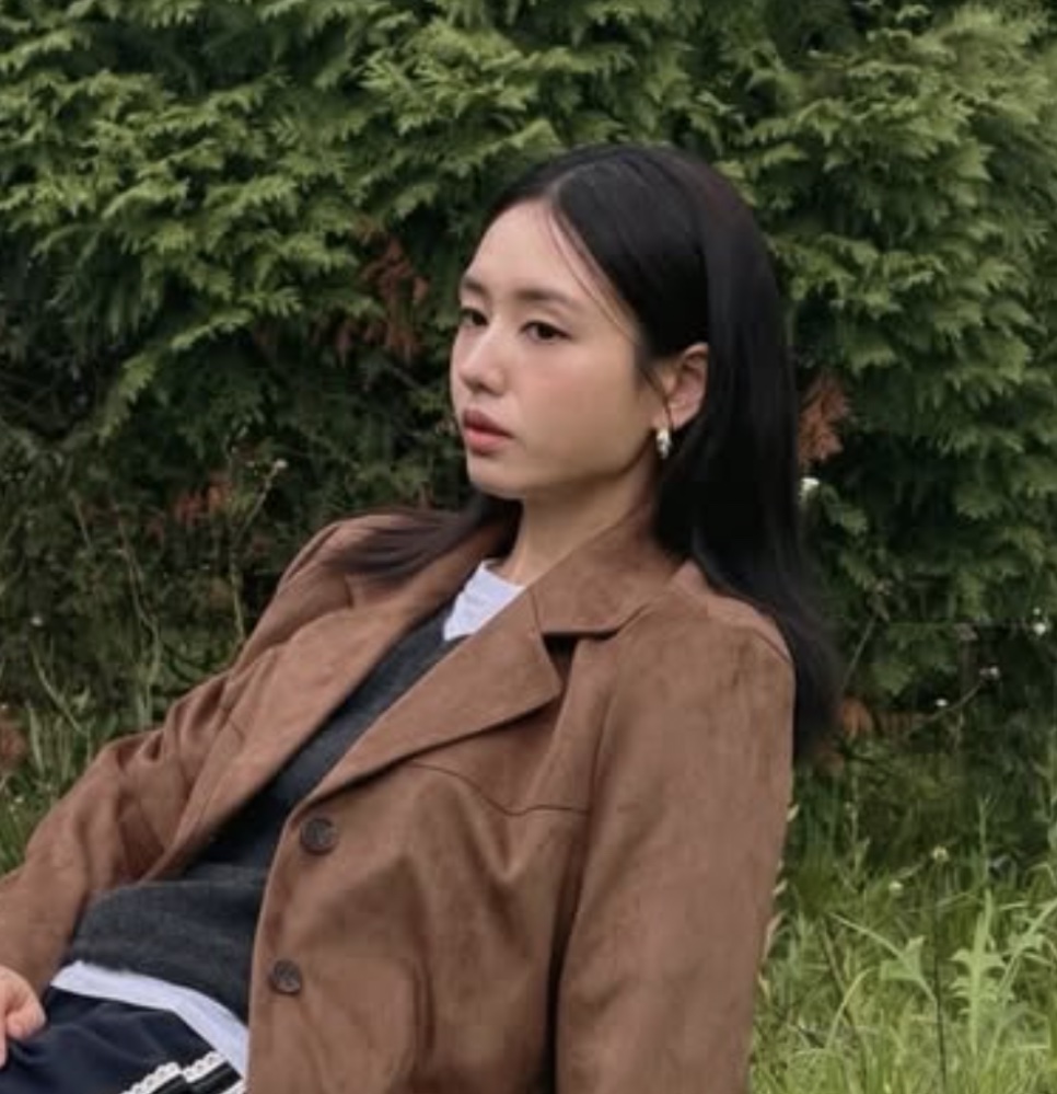Ahn Eun-jin de jaqueta marrom, sentada na grama na frente de uma cerca viva