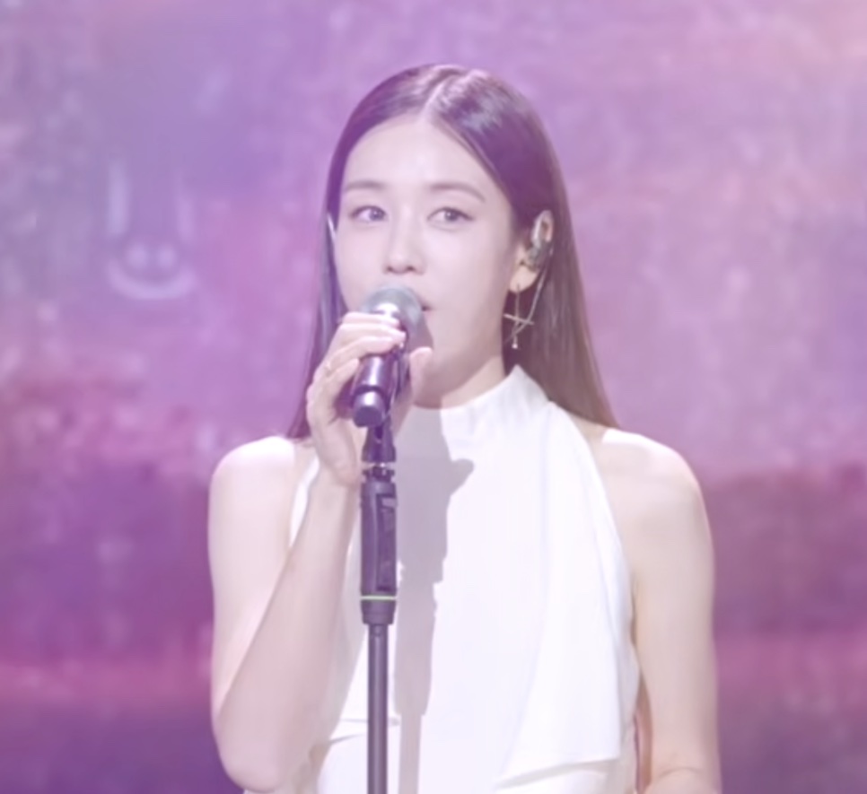Ahn Eun-jin de blusa branca, cantando no microfone em um palco com luz roxa