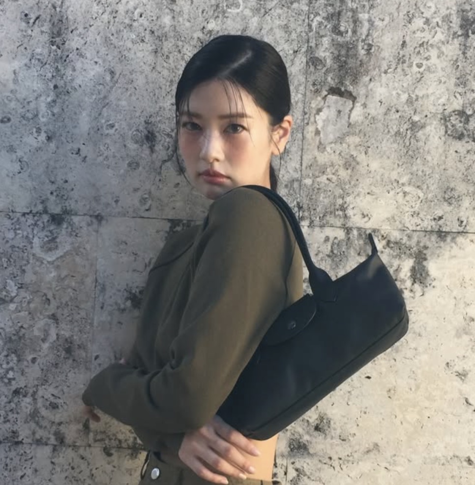 A atriz Jung So-min de jaqueta verde e bolsa preta no ombro, encostada em uma parede de pedra