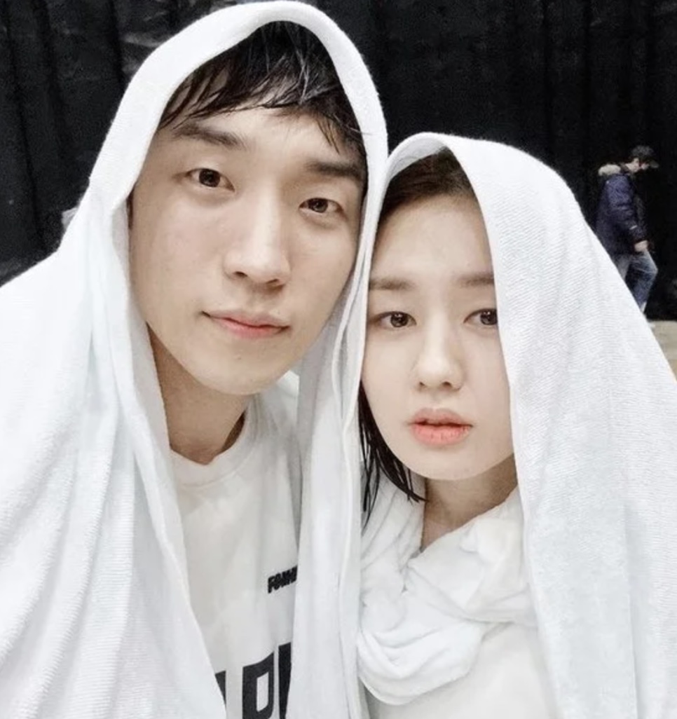 Ahn Eun-jin e um colega posando juntos para uma selfie, os dois com toalhas brancas na cabeça