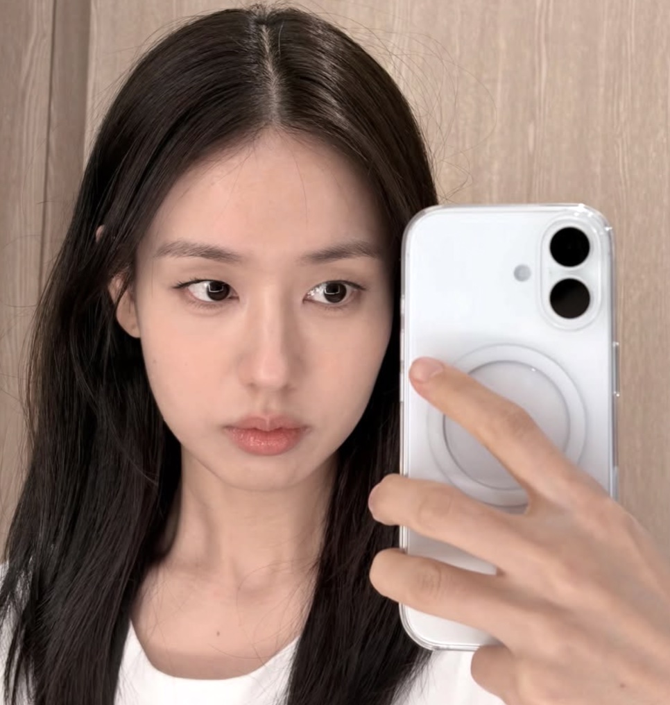Ahn Eun-jin de cabelo solto, tirando selfie no espelho com um celular branco