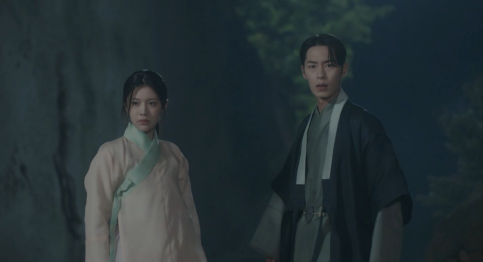 Jo-yeong e Jang Uk de pé diante de um paredão de pedra, à noite