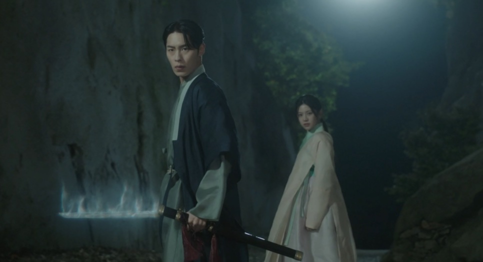 Jang Uk segurando a espada perto de chamas azuis, com Jo-yeong atrás dele, diante de um paredão de pedra