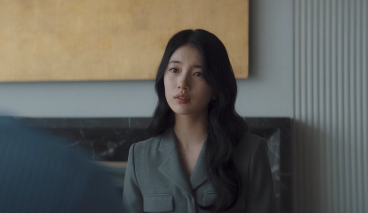 Suzy de blazer cinza, sentada em um quarto de hotel