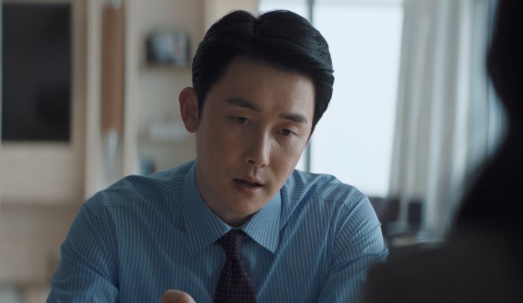 Kim Jun-han de camisa azul e gravata, sentado em uma sala de reunião
