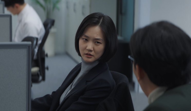 Park Ye-young de blazer escuro, virada para trás em uma redação, conversando com um colega
