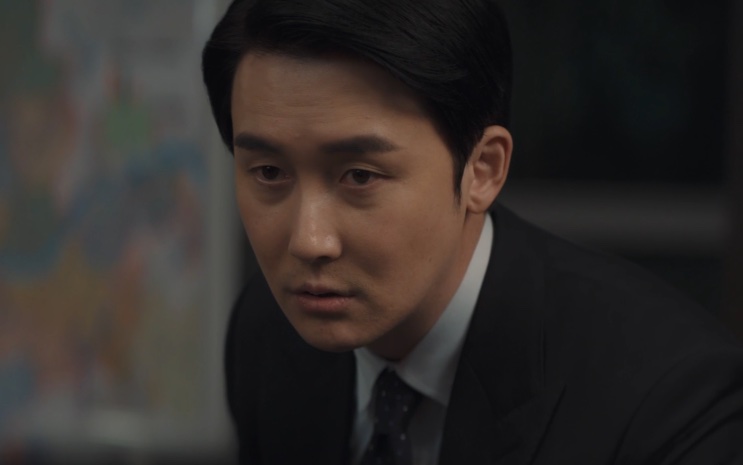 Choi Ji-hoon de terno, sentado e olhando para o lado