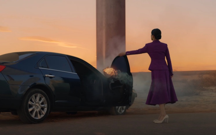 Carro em chamas em uma estrada ao pôr do sol, com Lee Yu-mi de pé ao lado