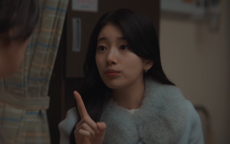 Lee Yu-mi de casaco cinza, com o dedo levantado, falando dentro de uma casa