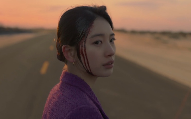 Lee Yu-mi de casaco roxo em uma estrada deserta, com o rosto marcado de sangue