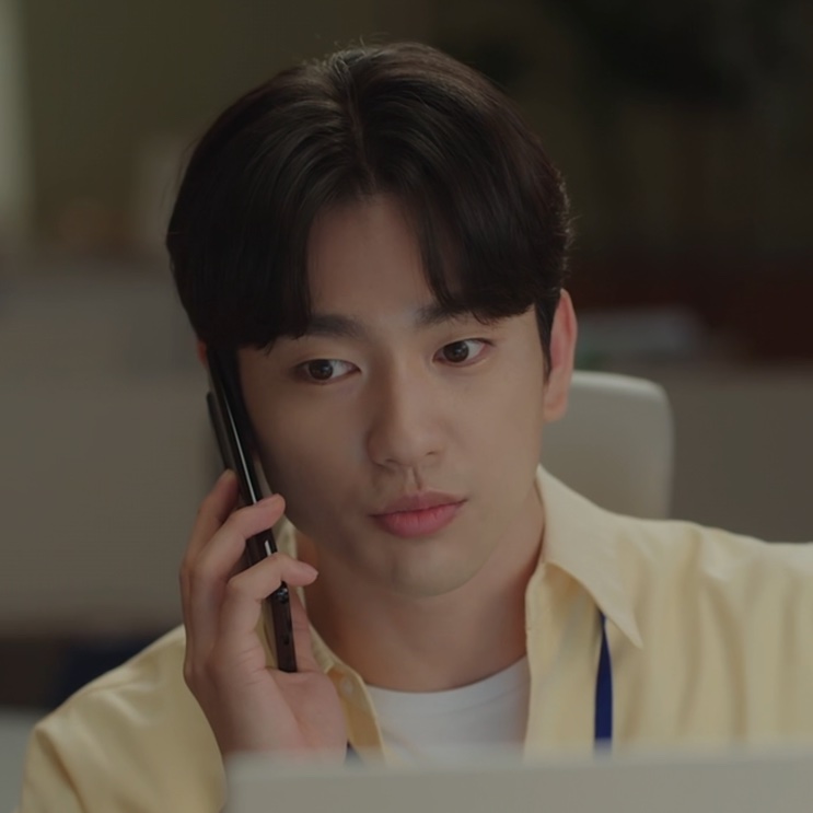 Yoo Babi de camisa amarela, falando ao celular na mesa do escritório
